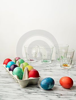 Colored Easter eggs in carton box on kitchen counter, emply glasses wirh remnants of dye