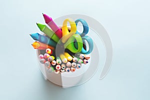 Colored pencils, scissors, notebook, ruler, pen, eraser, sharpener and more in glass. School and office stationery on light blue