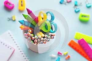 Colored pencils, scissors, notebook, ruler, pen, eraser, sharpener and more in glass. School and office stationery on light blue