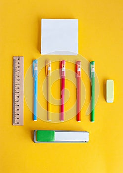 Colored pens, next to a wooden ruler, eraser, stapler, paper for notes, accessories for studying on a yellow background