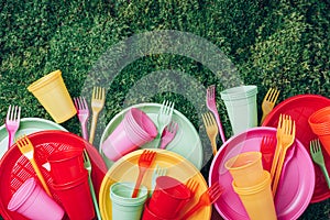 Colored plastic disposable tableware on green grass moss background. Top view. Copy space. Birthday picnic utensil