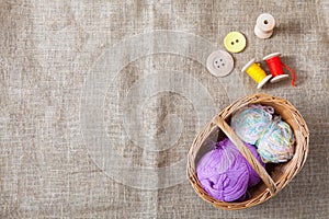 Colored threads and wooden buttons and a wicker basket are on sackcloth in the corner of the frame