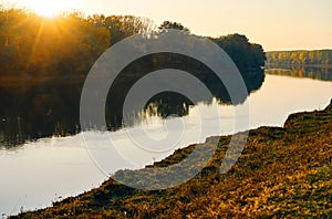 Colorful autumn forest landscape at sunset, trees near river and sky, bright sunlight reflected in water