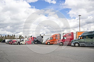 Colorful big rigs semi trucks with different semi trailers standing in row on truck stop parking lot taking break for truck