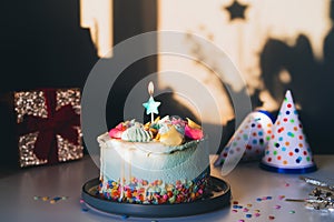 Colorful birthday cake with sprinkles and star shaped candle, festive caps and gift box on a dark wall background with