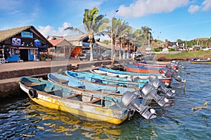 Colorful boats in the port of Hanga Roa, near Plaza Hotu Matua, surrounded by palm trees, against a blue sky.