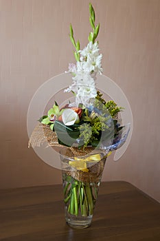 Colorful bouquet with such flowers as gladiolus, orchid, roses, callas, mimosa are standing in the big glasses vase. Tints of whit