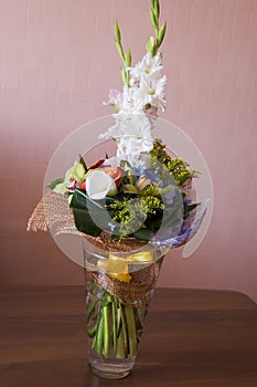 Colorful bouquet with such flowers as gladiolus, orchid, roses, callas, mimosa are standing in the big glasses vase. Tints of whit