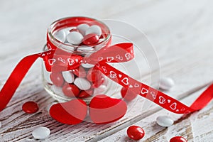 Colorful candy jar decorated with a red bow with hearts on white wooden background. Valentines day concept