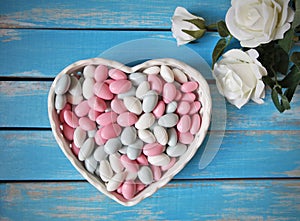 Colorful candy in white heart shaped bowl and white roses on wooden table