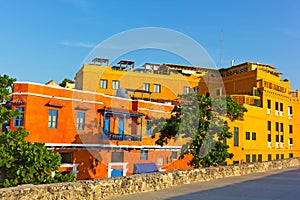 Colorful Cartagena walled city architecture brightly lit by sunset sunrays and surrounded by evergreen tropical plants.