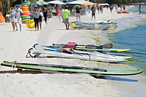 Colorful clear-bottomed rowing boats and surfboards on Boracay's sandy beach with palm trees and people in the background