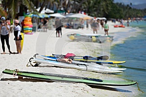 Colorful clear-bottomed rowing boats and surfboards on Boracay's sandy beach with palm trees and people in the background