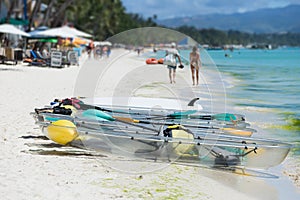 Colorful clear-bottomed rowing boats and surfboards on Boracay's sandy beach with palm trees and people in the background