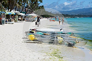 Colorful clear-bottomed rowing boats and surfboards on Boracay's sandy beach with palm trees and people in the background