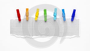 Colorful Clothespins Lined Up to a Torn Piece of White Copier Paper Against a Plain Background