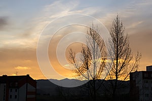 Barevné mraky nad městem a budovami nebo parkem a loukou, při východu nebo západu slunce.