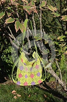 Colorful crocheted handbag with green handle behind the bush in the garden in summer