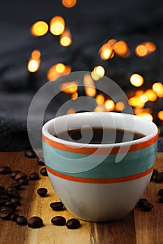 Colorful cup of black coffee with a cozy warm blanket on a wooden surface with the coffee beans and bokeh lights in the background