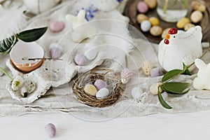 Colorful easter chocolate eggs in nest, spring flowers, chicken figurine and linen cloth on rustic wooden table. Space for text.