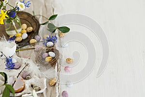 Colorful easter chocolate eggs in nest, spring flowers, feathers and linen cloth on rustic wooden table. Space for text. Easter