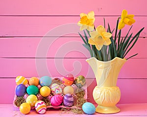 Colorful Easter Eggs in Carton with Vintage Yellow Vase filled with Spring Daffodils against Bright Pink Wood Board Background wit