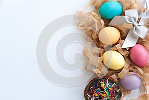 Colorful easter eggs on hay, easter cake and silver bow on white background flat lay. Copy space. Top view