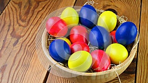 Colorful easter eggs lie in a plate on a straw on a wooden table.