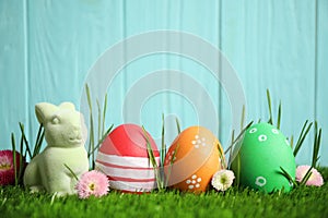 Colorful Easter eggs, rabbit and daisy flowers in green grass against blue background. Space for text