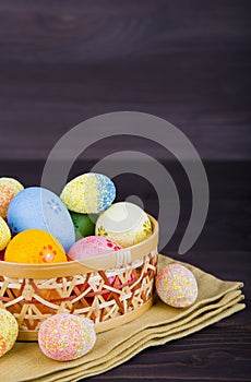 Colorful Easter eggs in wicker basket on textile on dark wooden