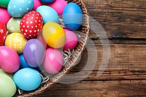 Colorful Easter eggs in wicker basket on wooden table, top view. Space for text