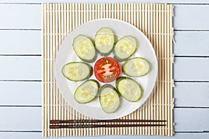 Colorful fresh dietary snack from natural vegetables on white plate. Sliced red tomato, cucumber in form of flower and chopsticks.