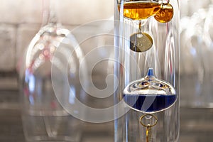 A colorful, glass, liquid filled Galileo Thermometer measures indoor room temperature in a wine cellar with wine glasses hanging