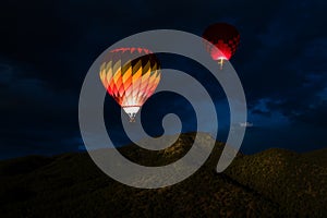 Colorful glowing hot air balloons floating in a night sky over forested mountain peaks