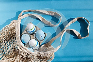 colorful handmade painted easter eggs in a string bag on blue background with hard shadows.