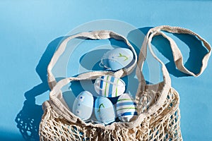 colorful handmade painted easter eggs in a string bag on blue background with hard shadows.