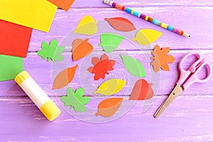 Colorful leaves cut from colored paper, scissors, glue stick, pencil, paper sheets on purple wooden background. Top view