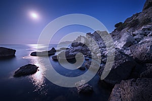 Colorful night landscape with full moon, lunar path and rocks in summer. Mountain landscape at the sea.