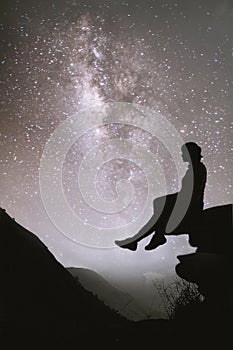 Colorful night sky with stars and silhouette of a standing girl sitting on the stone. Blue milky way with girl on the mountain.