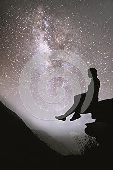 Colorful night sky with stars and silhouette of a standing girl sitting on the stone. Blue milky way with girl on the mountain.