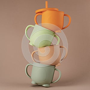 Colorful pastel silicone sippy cups stacked in column on brown background. Baby tableware, feeding concept.