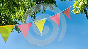 colorful pennant string decoration in green tree foliage on blue sky, summer party background