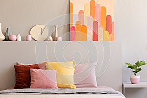 Colorful pillows on bed in grey bedroom interior with poster and clock on bedhead. Real photo