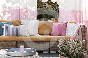 Colorful pillows on rattan sofa on the terrace with flowers, painted drapes and candles. Real photo