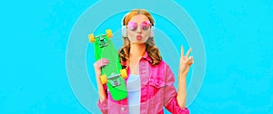 Colorful portrait of stylish young woman with skateboard listening to music in headphones wearing pink jacket, sunglasses on blue