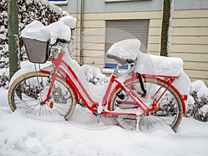 A colorful red bike covered with many fresh snow standing in front of a house facade.
