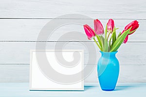 Colorful red spring tulip flowers in nice blue vase and blank photo frame on light wooden background as greeting card. Mothersday