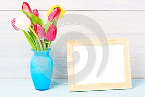 Colorful red spring tulip flowers in nice blue vase and blank photo frame on light wooden background as greeting card. Mothersday