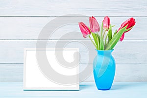 Colorful red spring tulip flowers in nice blue vase and blank photo frame on light wooden background as greeting card. Mothersday