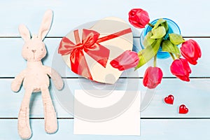 Colorful red spring tulip flowers in nice blue vase, blank photo frame and stuffed toy bunny with decorative heart on light wooden